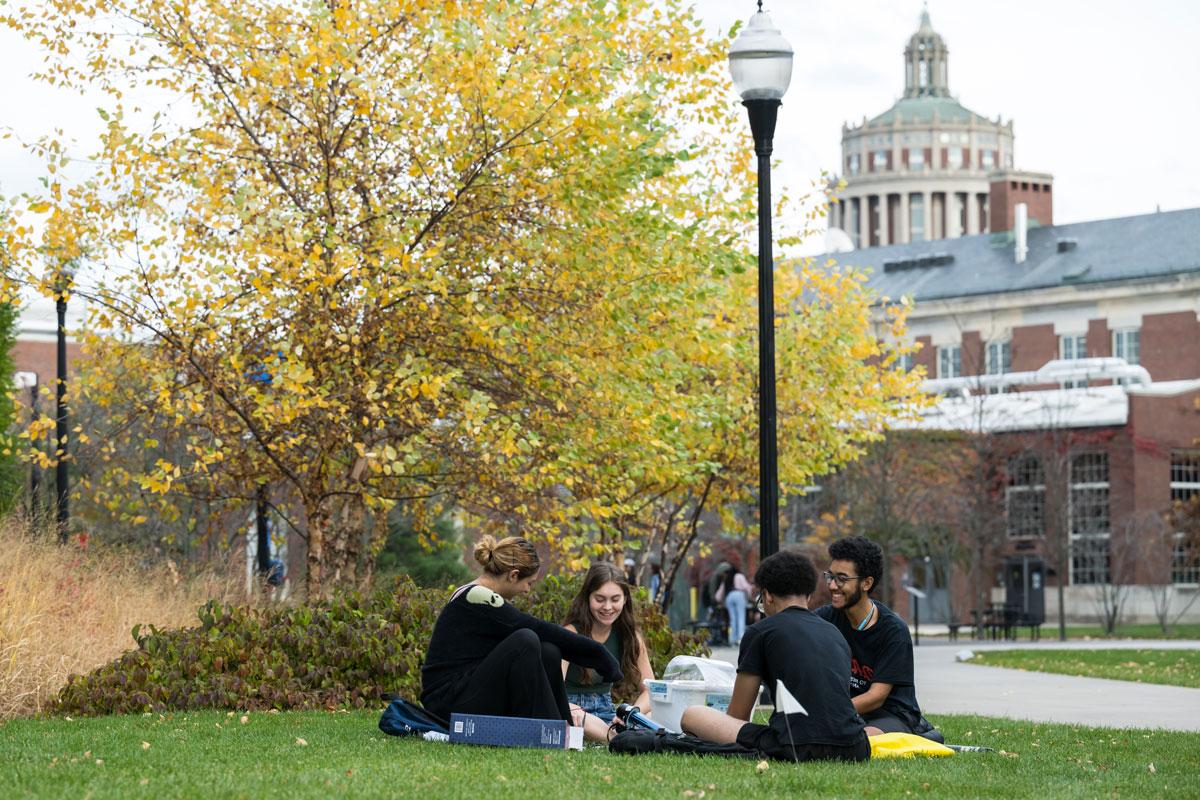 A group of students sitting on the grass at the University of Rochester campus, enjoying a sunny day together.